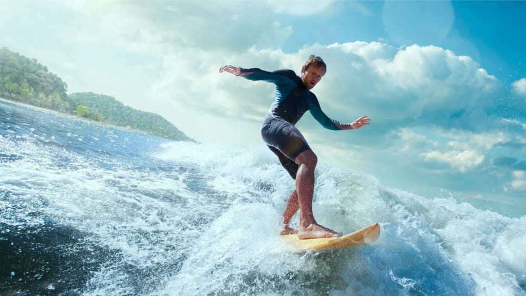A man in a wetsuit surfs on a wave near the shore under a partly cloudy blue sky.